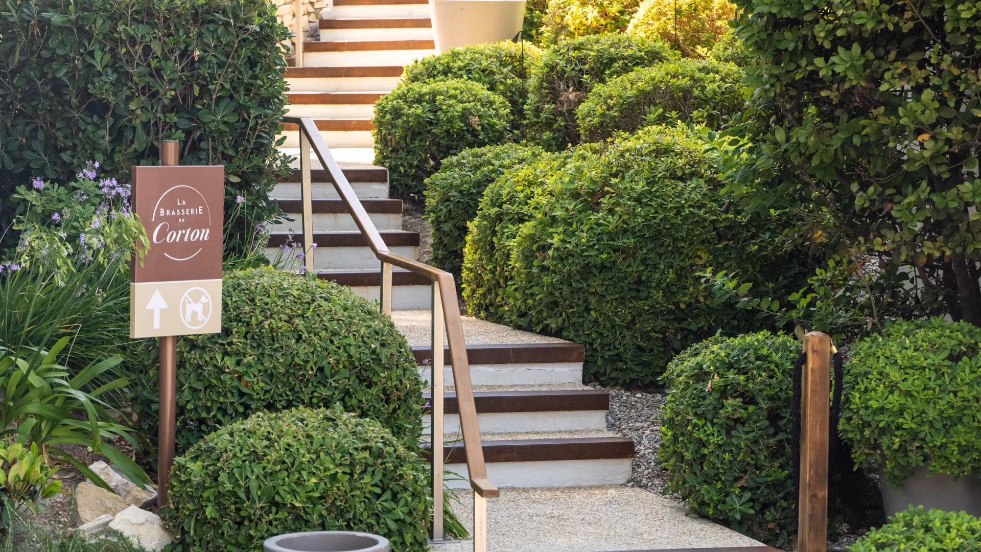 Un escalier en pierre avec des rampes en bois mène à une terrasse arborée, accompagné d'un panneau indiquant "La Brasserie Corom"