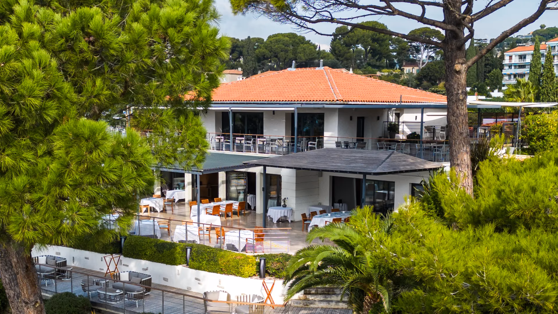 Restaurant avec terrasse et tables en extérieur, entouré d'arbres et de végétation, sous un ciel partiellement nuageux